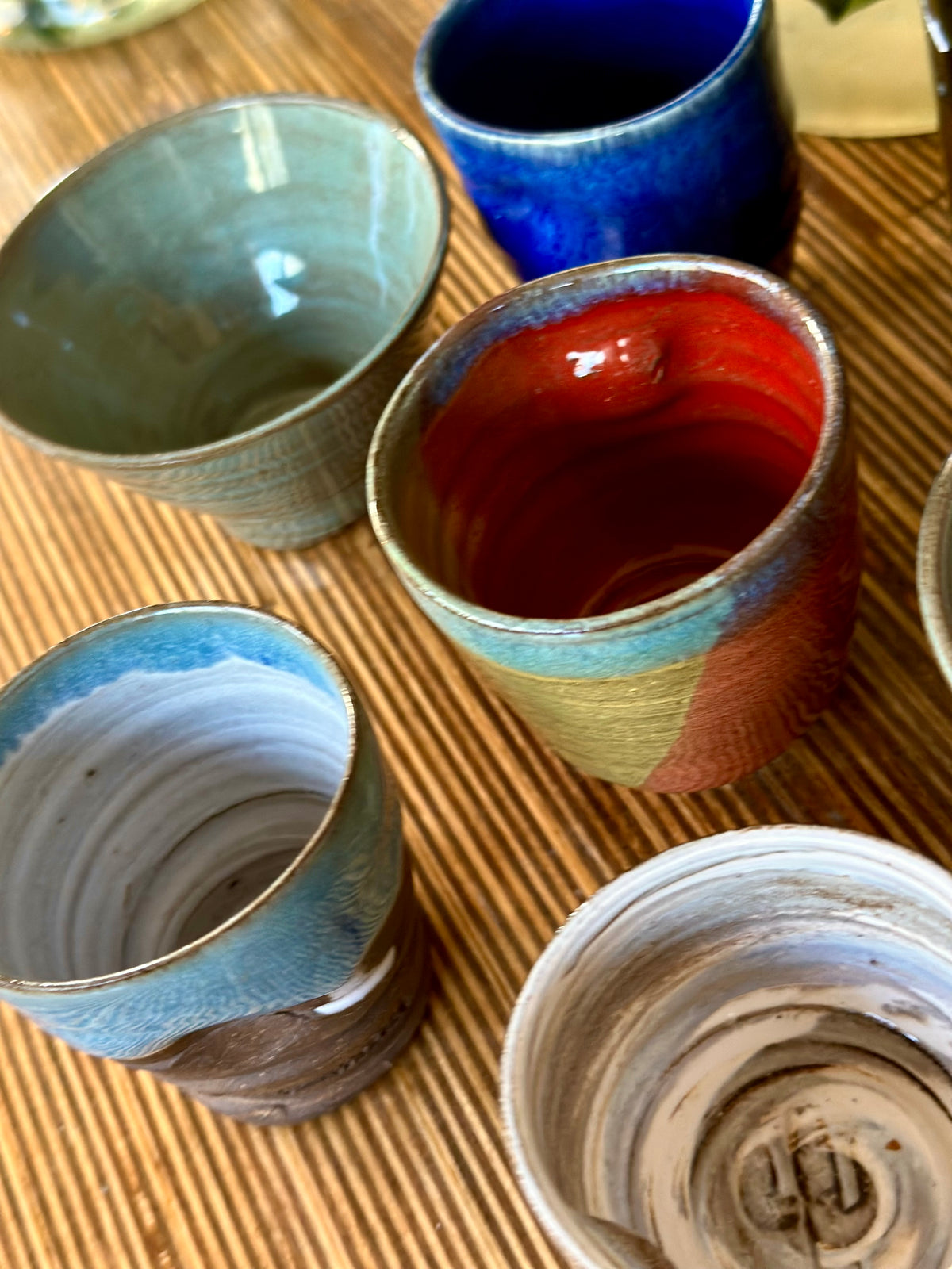 A group of handmade ceramic cups with various glazed finishes, including blue, red, beige, and white swirls, arranged on a woven straw mat.
