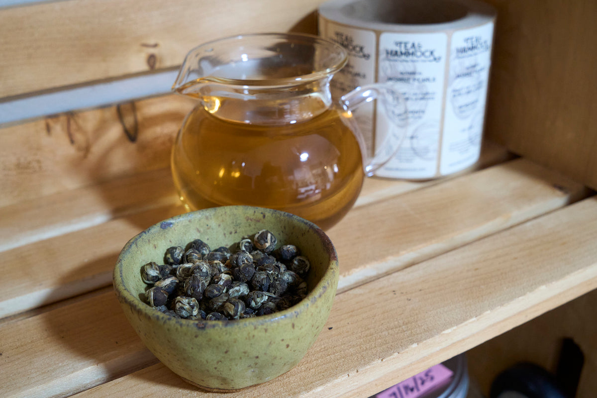 tea & hammock jasmine pearls tea displayed in beautiful tea bowl with vibrant brewed tea in clear teapot in background displayed in wooden crate