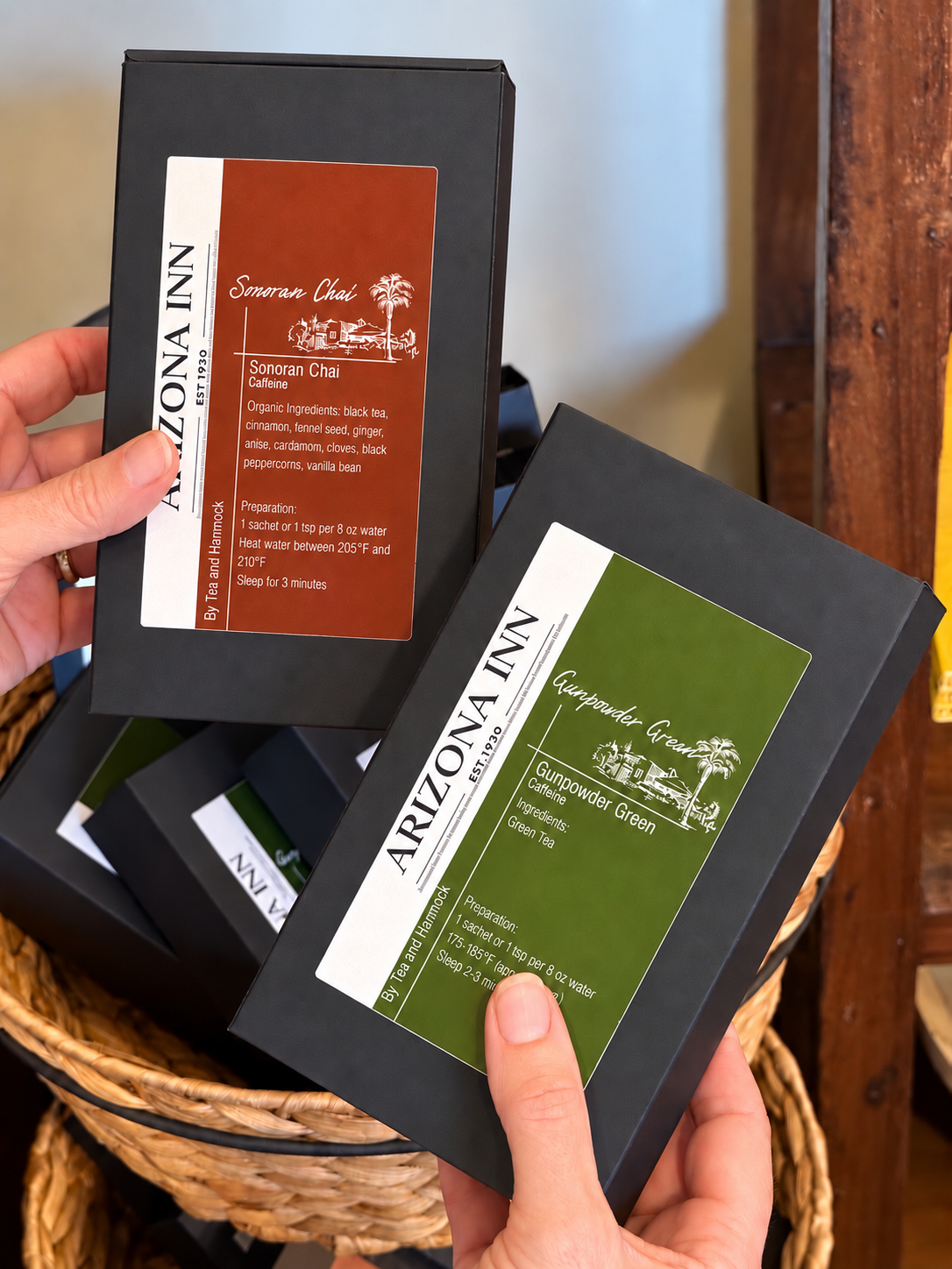 Two Arizona Inn Coffee Company black cardboard boxes held in hands over a woven basket. One box is labeled 'Sour Cream Cafe' in brown, and the other is labeled 'Cappuccino Cream' in green.