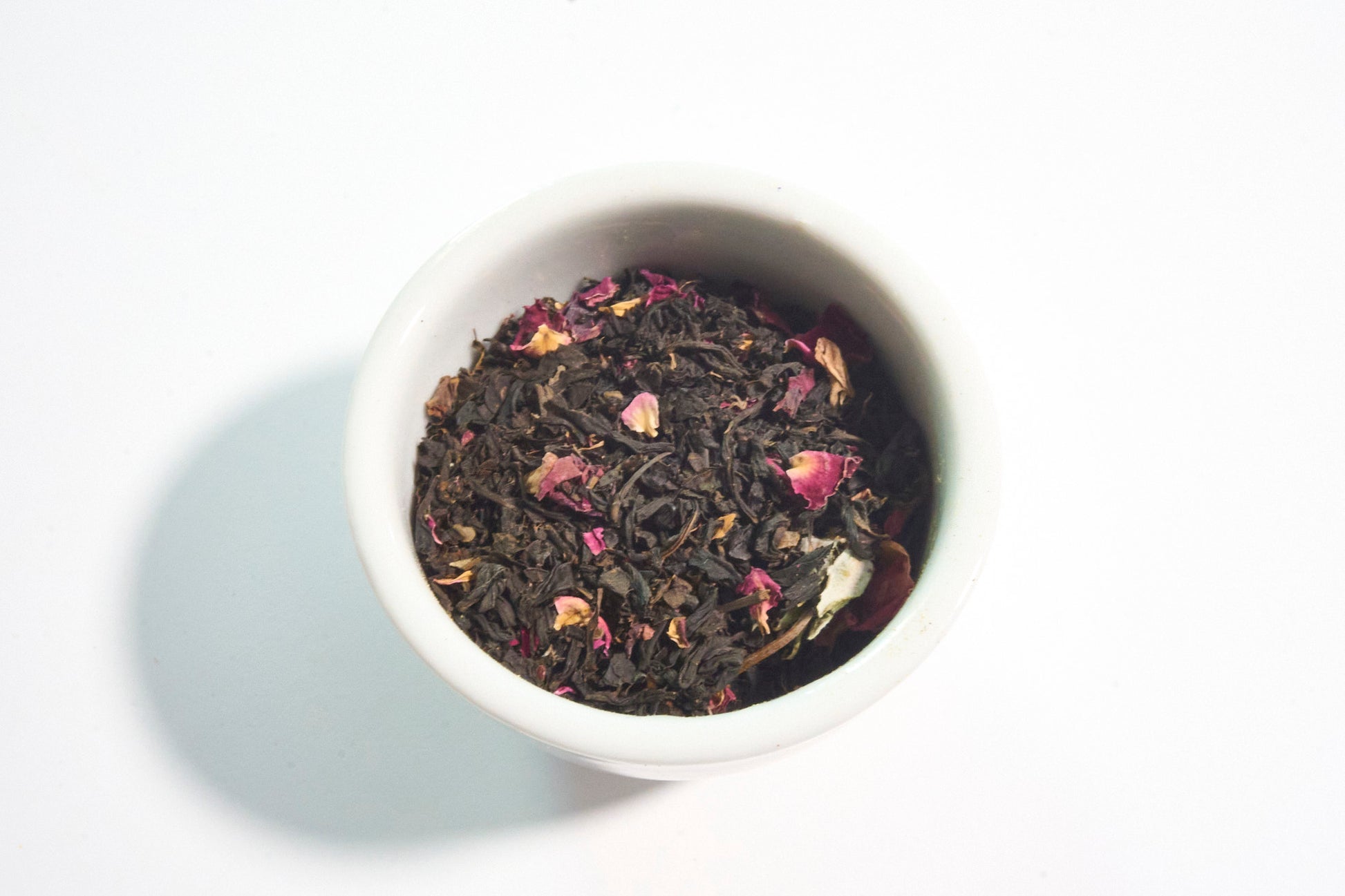 White bowl filled with black tea leaves and pink flowers on a white background