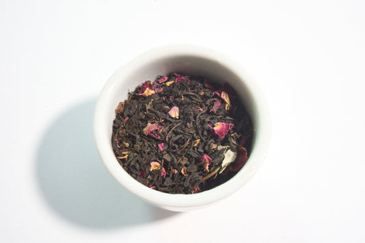 White bowl filled with black tea leaves and pink flowers on a white background