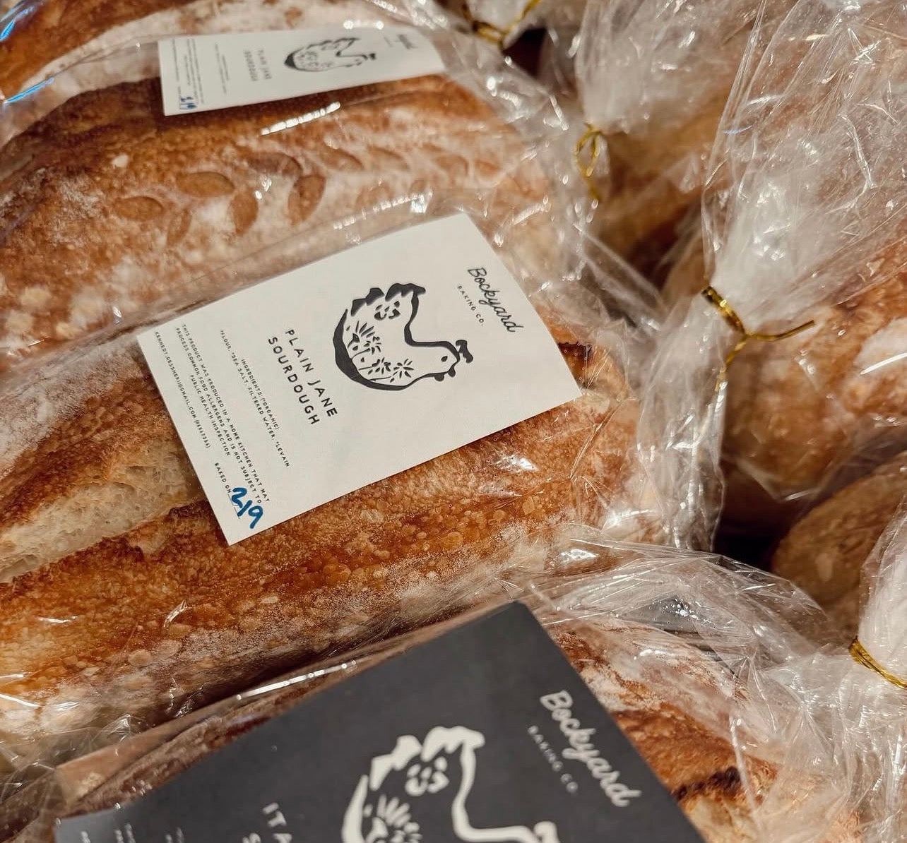 Several loaves of crusty artisan bread wrapped in clear plastic bags with white labels reading Backyard Farming, stacked together.