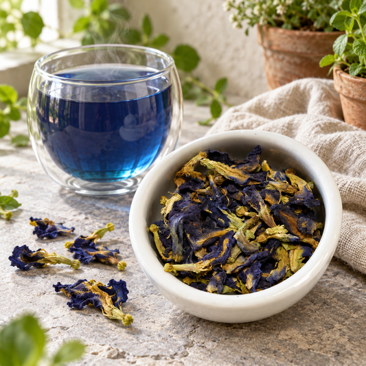A white ceramic bowl filled with dried blue butterfly pea flowers, next to a clear glass cup of vibrant blue tea, on a stone surface with green plants in the background.