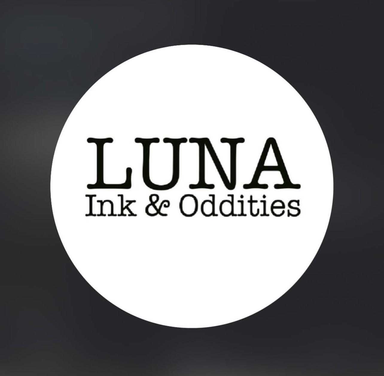 A white circular badge or sticker with the text 'LUNA Ink & Oddities' in black serif font, centered on a dark gray gradient background.