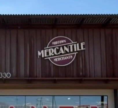 A storefront sign reading 'Midtown Mercantile Merchants' mounted on a dark brown corrugated metal wall above a glass window.