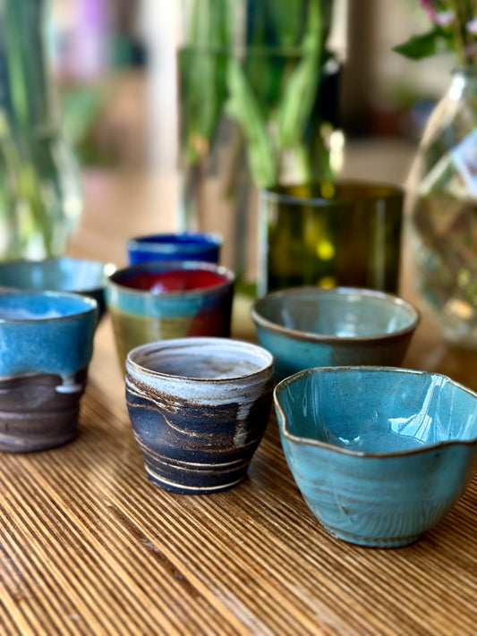 A group of handmade ceramic cups and bowls in various earthy glazes, including blue, teal, brown, and red, arranged on a bamboo mat with blurred glass vases in the background.