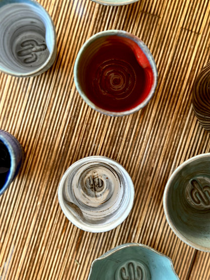 A top-down view of several handmade ceramic cups arranged on a woven straw mat. The cups have varied glazes in gray, white, blue, and a deep reddish-brown, with one cup filled with dark red liquid.