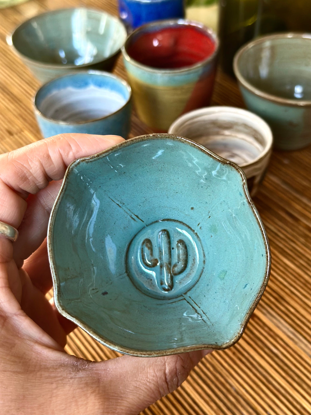 A handmade ceramic cup with a scalloped rim and an embossed cactus motif in the center, held by a hand over a bamboo mat background with other colorful cups.
