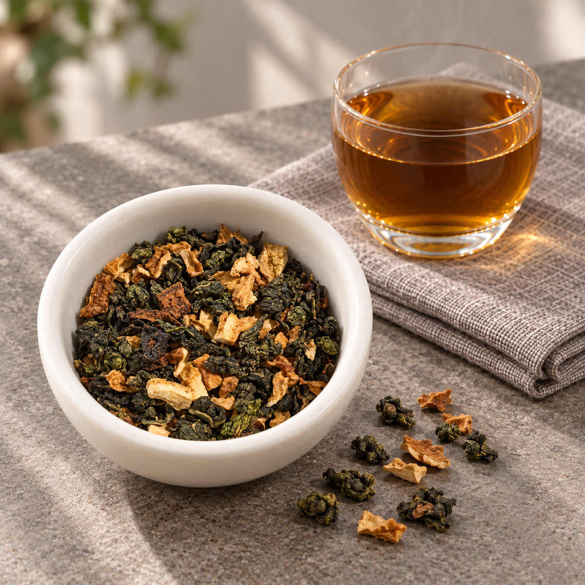 A white bowl filled with Toasted Orange Oolong loose-leaf tea, with a glass of brewed amber tea and scattered tea leaves on a textured gray surface.