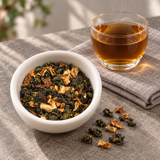 A white bowl filled with Toasted Orange Oolong loose-leaf tea, with a glass of brewed amber tea and scattered tea leaves on a textured gray surface.