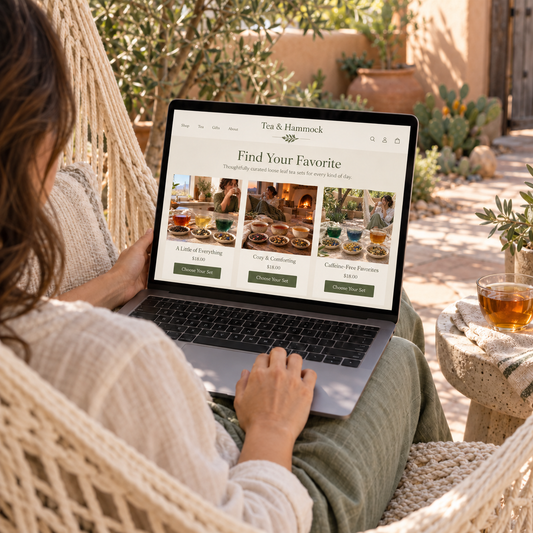A woman relaxes in a cream macrame hammock outdoors, using a laptop to browse the Tea & Hammock website. The scene is set in a sunny, tranquil patio garden with a small stone side table holding a glass of tea nearby.