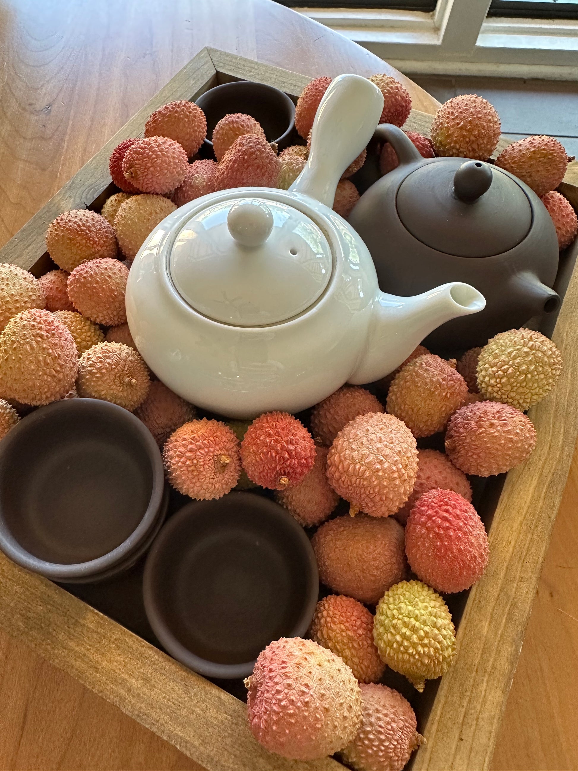 tea & hammock lychees for lychee tea with white japanese style teapot and brown teapot with matching teacups. the teaware is displayed in a crate of lychees